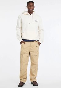 Pantalon cargo beige avec plusieurs poches, associé à un sweat à capuche crème avec capuche et cordons. La tenue est complétée par des chaussures marron.