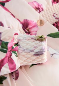Tissu matelassé rose pastel avec des motifs floraux et de feuilles, montrant un poignet et un petit bouton rond sous un éclairage doux.