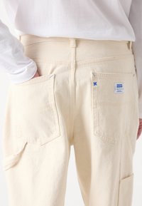 Pantalons en denim couleur crème avec deux poches à l'arrière, des détails de couture subtils et une étiquette portant la mention "HUGO" près de la taille.