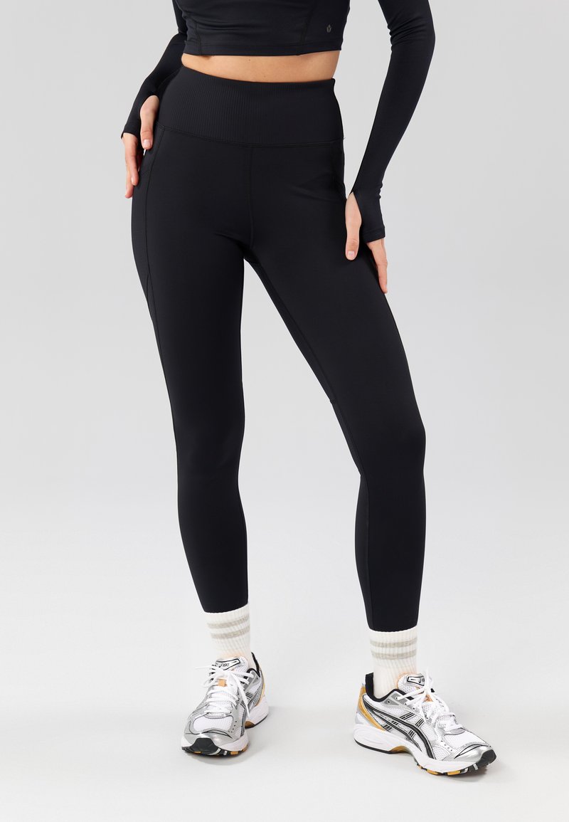 Leggings noirs taille haute en tissu lisse et extensible avec des détails de coutures. Assortis à des chaussures de course argentées et des chaussettes blanches à rayures.