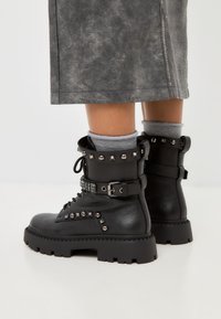 Botas de cuero negras hasta el tobillo con acentos de tachuelas, diseño de cordones y suelas gruesas. Cuenta con un acabado texturizado y hebillas decorativas.