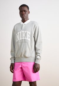 Siv sweatshirt z logotipom "NIKE", kengurska luknja in ribani manšeti, v kombinaciji z roza kratkimi hlačami. Bel ovratnik srajce pokuka iz izreza.