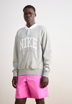 Grijze sweatshirt met het "NIKE" logo, buidelzak en ribgebreide manchetten, gecombineerd met roze shorts. Een witte overhemdkraag steekt uit de halslijn.