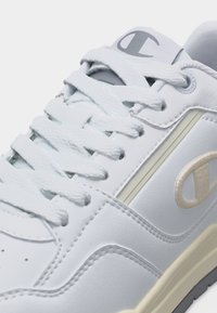 Άσπρο δερμάτινο αθλητικό παπούτσι με λευκά κορδόνια, το λογότυπο Champion στη γλώσσα και στην πλευρά, και ανοιχτό μπεζ σόλα ορατή στη δεξιά άκρη.