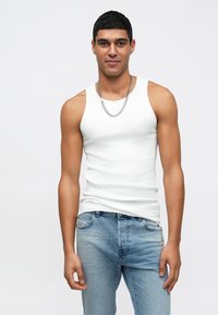 Homem com cabelo preto curto, vestindo uma camisola branca sem mangas, colar de corrente prateada e jeans de tom azul claro, posando contra um fundo branco simples.