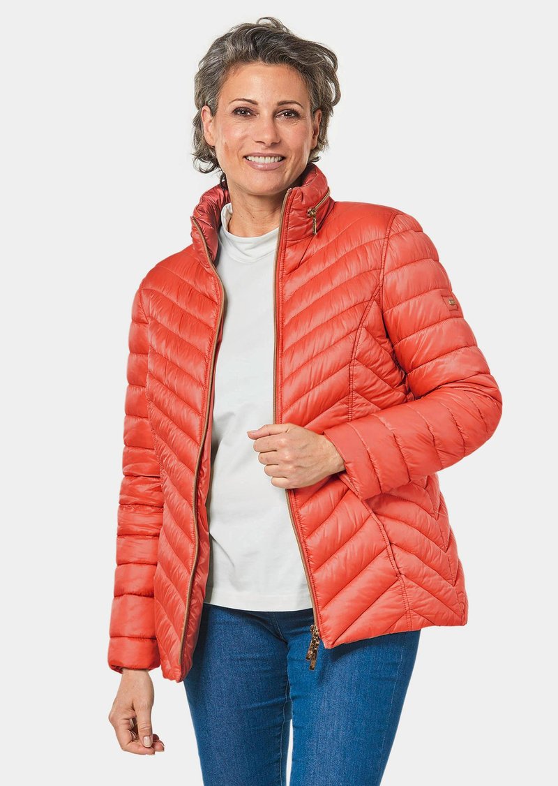 Padded orange jacket mit einem Reißverschluss, Chevron-Steppmuster, Stehkragen und leichtem Material. Über einem weißen Hemd getragen.