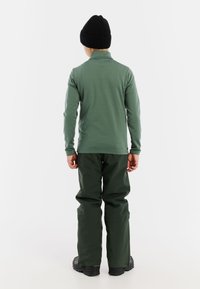 Camicia verde a maniche lunghe e pantaloni verdi, abbinati a un berretto di maglia nero. Il materiale appare morbido e alla moda, con una vestibilità casual.