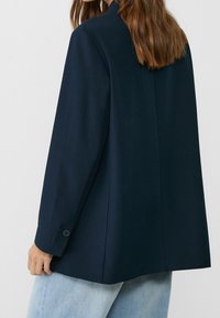 Blazer azul marino hecho de una tela suave, con un corte entallado, puños abotonados y un diseño liso en la parte posterior, combinado con jeans de color azul claro.