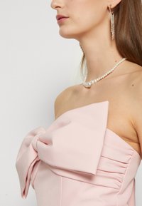 Abito rosa senza spalline con un grande fiocco decorativo e una texture arricciata sui lati. Accessoriato con una collana di perle e orecchini pendenti.