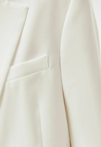 Närbild av en vit kavaj som visar den skräddarsydda slagkragen och en passpoalficka på vänster sida.