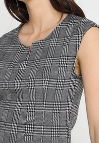 Haut sans manches à motif à carreaux noir et blanc, arborant un col rond et un tissu texturé subtil avec une coupe propre et ajustée.