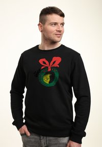 Černá mikina s grafikou zeleného věnce, na které se nachází hlava žluté kreslené postavičky, červená mašle a nápis "The Grinch."