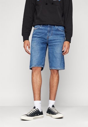 Mann trägt knielange blaue Jeansshorts, schwarzen Hoodie, weiße Socken und schwarze flache Sneaker, steht vor einem einfarbigen Hintergrund.