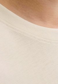 Nahaufnahme eines beigen Rundhalsstoffs auf einem Kleidungsstück, der gegen die sichtbare Haut am Ausschnitt wirkt.