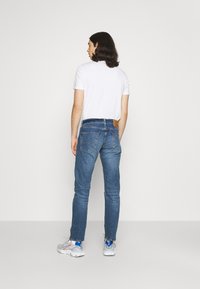 Jeans de mezclilla azul con corte recto, bolsillos traseros visibles, combinados con una camiseta blanca y zapatillas grises. Textura robusta, acentos mínimos.