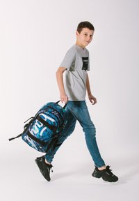 Zaino blu e nero con motivo e dettagli bianchi, realizzato in materiale resistente. Modello con una maglietta grigia e jeans blu che lo porta.