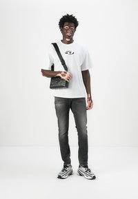 Camiseta blanca de algodón con logo, bolso cruzado negro, jeans gris deslavado y zapatillas en blanco y negro, de pie contra un fondo blanco.