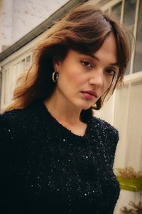 Õlgadeni ulatuvate pruunide juustega naine, kes kannab musta litritega topi ja hõbedast rõngakujulisi kõrvarõngaid, seisab hoone akende lähedal.