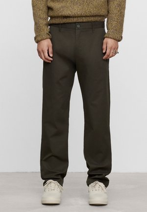 Persona che indossa pantaloni dritti color oliva scuro, maglione beige in maglia e scarpe da ginnastica bianche, in piedi davanti a uno sfondo semplice.