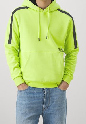 Person iført en neongrøn hættetrøje med sorte skulderstriber og blå jeans, hænderne i lommerne.