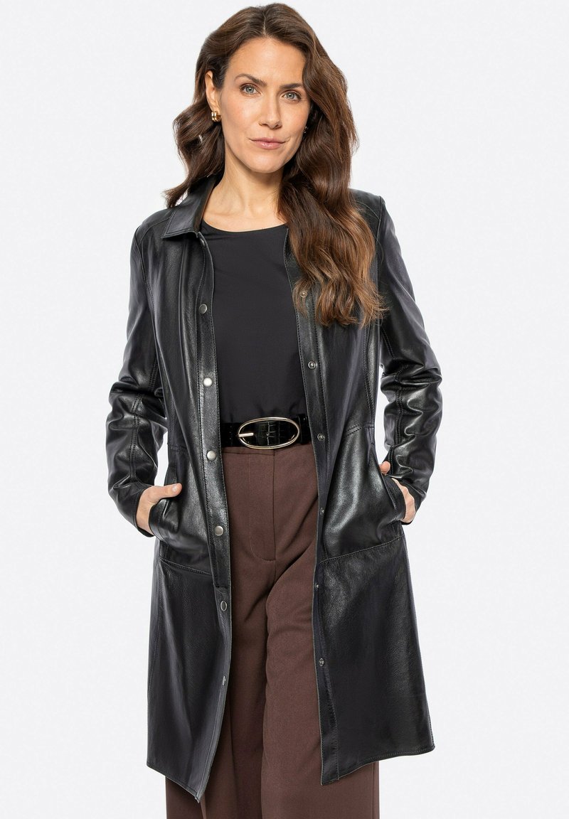 Abrigo de cuero negro con cierre de botones en la parte delantera, bolsillos y cuello. Textura suave y mangas largas, combinado con un cinturón y pantalones marrones.