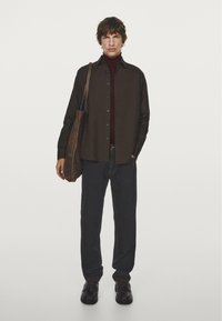 Chemise boutonnée à manches longues marron foncé sur un col roulé bordeaux, associée à un pantalon sombre et des chaussures à lacets marron. Sac à bandoulière marron.