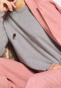 Sudadera gris con un escote redondo, combinada con una chaqueta texturada rosa. Un collar simple con un colgante de oro añade un toque de detalle.