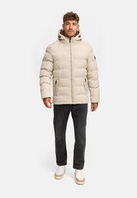 Doudoune beige avec capuche, présentant des sections matelassées verticales, une fermeture éclair à l'avant et deux poches latérales. Portée sur un jean foncé et des baskets.