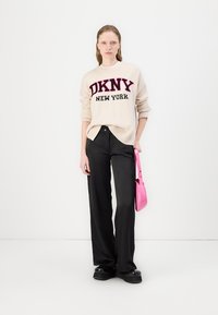 Pulôver de malha creme com o logotipo da DKNY em preto e rosa, combinado com calças largas pretas e uma bolsa rosa. Sapatos plataforma pretos.