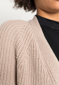Cardigan beige clair texturé avec motif côtelé et coupe ample. Porté sur un col roulé noir à manches longues.