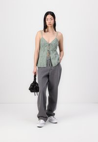 Svetlo zelena kvačkana bluza z V-izrezom in detajlom na vezanje, v kombinaciji s prostornimi sivimi hlačami. Črna torba z vzorcem in bele superge dopolnjujejo outfita.