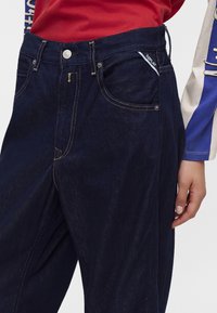 Personne portant un jean en denim bleu foncé avec des coutures jaunes et un haut rouge, main reposant à côté.