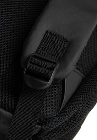 Dettaglio di uno zaino in rete nera con una robusta cinghia di regolazione dotata di una fibbia in plastica rotonda, accenti in tessuto liscio e una finitura texturizzata.