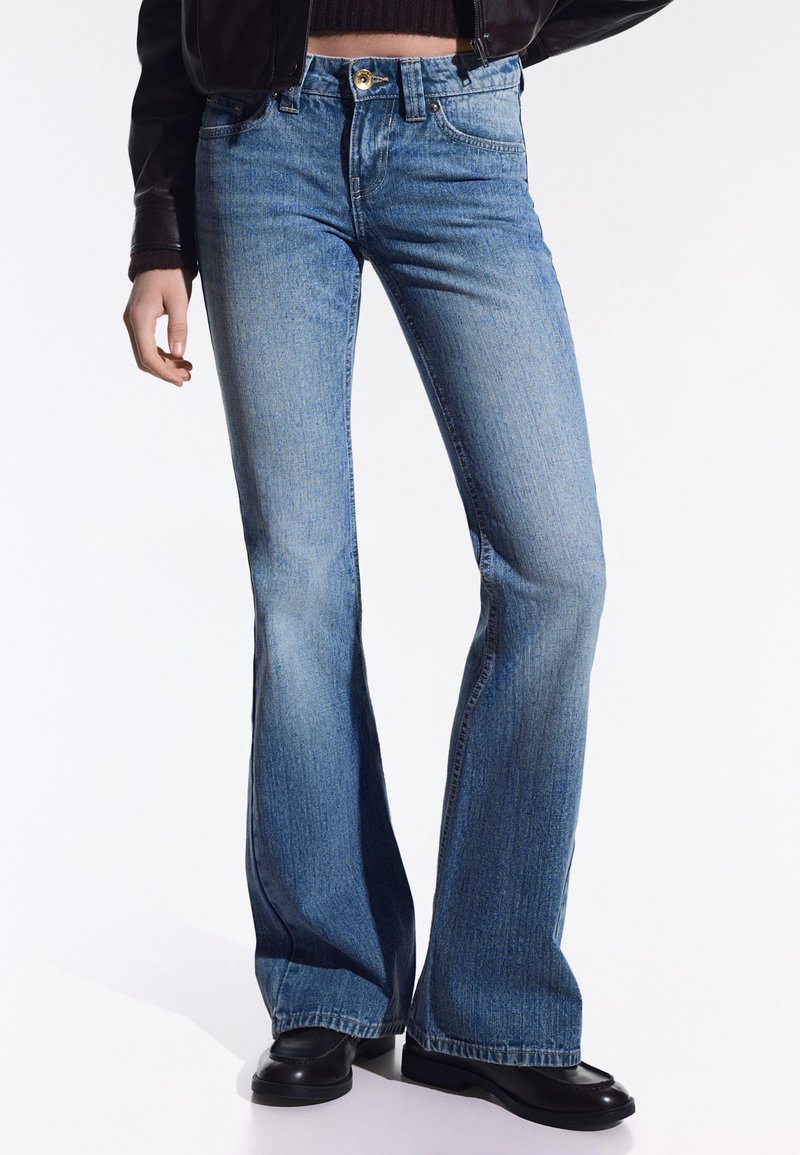 Personne portant un jean évasé bleu clair, une veste en cuir noire et des chaussures noires, debout devant un fond blanc.