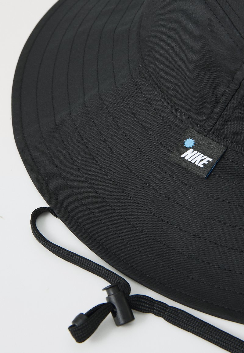 Sombrero de cubo negro hecho de tela, con un ala ancha y superficie texturizada. Incluye una etiqueta de Nike y una correa ajustable para la barbilla.