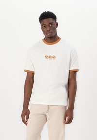 Camiseta blanca de algodón con un cuello y puños de manga acanalados en marrón. Presenta un diseño impreso de tres ranas estilizadas en el pecho.