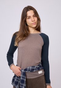 Maglietta a maniche lunghe di colore marrone chiaro con maniche blu navy; presenta un collo tondo e una camicia di flanella a quadri annodata intorno alla vita.