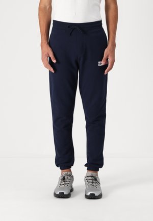 Mężczyzna ubrany w granatowe spodnie dresowe z elastycznymi mankietami i białą koszulę, stojący w szaro-czarnych butach sportowych na białym tle.