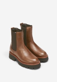 Paire de bottines Chelsea en cuir marron avec panneaux latéraux élastiques noirs et semelles épaisses cloutées noires sur fond blanc.