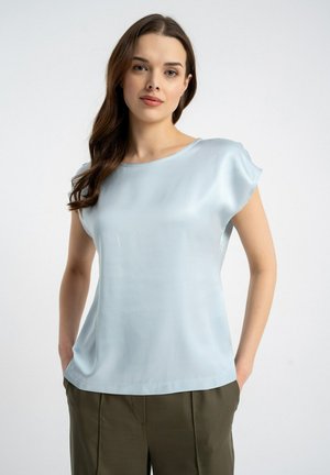 Kobieta o długich brązowych włosach, ubrana w jasnoniebieską satynową bluzkę z krótkim rękawem i oliwkowozielone spodnie, stoi z rękami w kieszeniach.