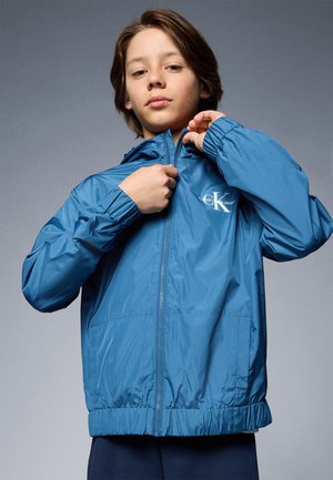 Giacca leggera blu con cappuccio, chiusura zip, polsini e vita elasticizzati. Presenta un logo sul petto e una texture liscia e lucida.