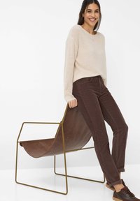 Giovane donna sorridente, appoggiata su una moderna sedia a sella in pelle marrone con telaio in metallo dorato, indossando un maglione beige e pantaloni marrone scuro.