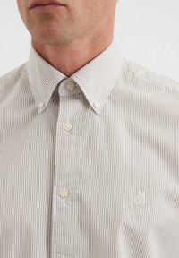 Gestreiftes Hemd in hellbeige und weiß mit einem Umlegekragen, Perlmuttknöpfen und einem kleinen Logo auf der Brust gestickt.