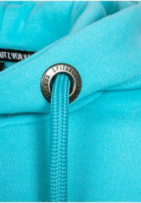 Turquoise hoodie close-up showing a braided drawstring and a round silver grommet with "SPITZBUB" engraved. Soft texture, minimal design details.