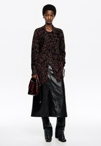 Cárdigan marrón con estampado de leopardo y cierre de botones, superpuesto a una falda de cuero oscura y pantalones a juego, acompañado de un bolso de cuero burdeos.