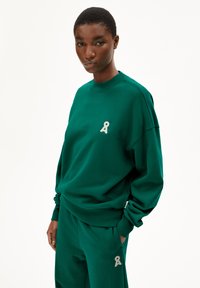 Grüner Baumwollpullover mit Rundhalsausschnitt, der ein kontrastierendes hellgraues Logo auf der Brust aufweist. Der Stoff wirkt strukturiert und weich.