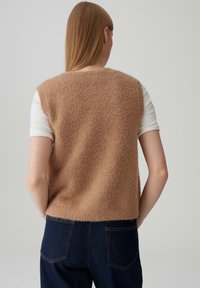 Gilet beige morbido con un design largo, abbinato a una camicia bianca e jeans in denim scuri. Il gilet ha una trama soffice e una silhouette corta.
