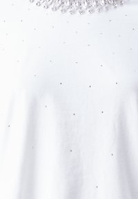 Valge t-särk sileda tekstuuriga, millel on väiksed säravad kristallid, mis on laiali jaotatud kangale, ning dekoratiivne rhinestone krae.