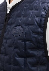 Μπλε ναυτικό γιλέκο με ραμμένη σχεδίαση, μαύρο φερμουάρ και επιγραφή λογότυπου Emporio Armani EA7 στο στήθος, φορεμένο πάνω από λευκό πουκάμισο.