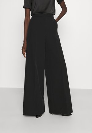 Persoon die zwarte, hoge taille, wijde broek draagt en een zwart, kort mouw shirt in de broek gestopt heeft, staand met één hand in de zak.